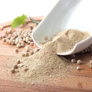 Finely ground White Pepper powder pouring from a white ceramic bowl onto a wooden cutting board.